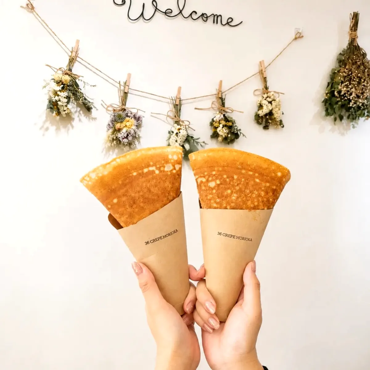 白壁にWelcomeのワイヤーサインとドライフラワーのガーランド。クラフト紙に包まれた36 CREPE MORIOKAのクレープを両手で掲げている